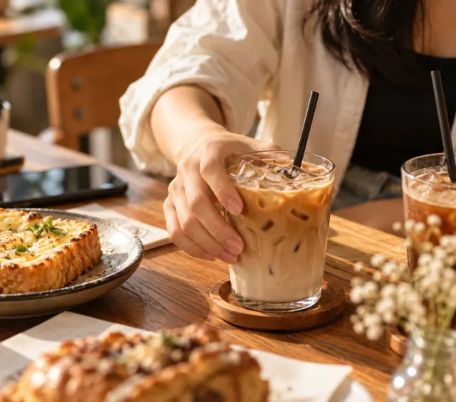 Tangan mengangkat segelas es kopi susu di atas meja kayu, di samping roti panggang.